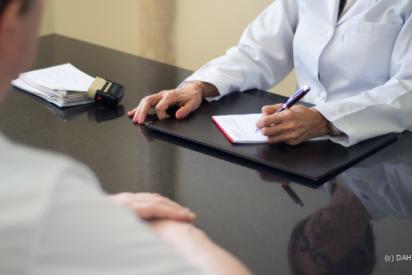 Ärztin sitzt an ihrem Schreibtisch und macht sich Notizen auf ihrem Block. Ihr gegenüber sitzt ein männlicher Patient.