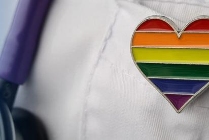 Anstecker in Form eines Herzens in Regenbogenfarben steckt an einem Arztkittel, daneben ein Stetoskop.