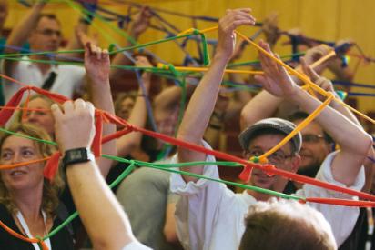Viele Menschen in einem Konferenzsaal halten an ausgestreckten Armen ein Netz aus zusammengeknüpften bunten Bändern in die Luft