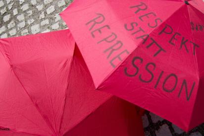 Zwei rote Regenschirme liegen aufgespannt auf Kopfsteinpflaster, auf dem rechten steht geschrieben: Respekt statt Repression
