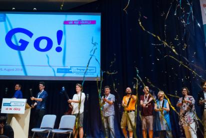 Auf einer Bühne stehen mehrere Menschen mit Konfetti-Kanonen, aus denen Girlanden geschossen werden. Im Hintergrund steht auf einer Projektionswand: Go!